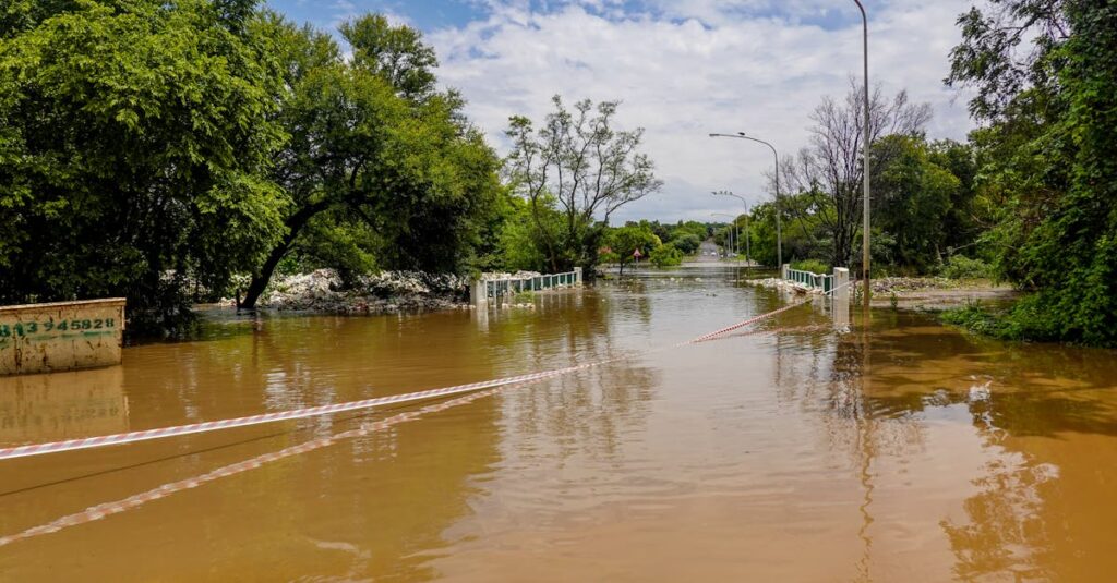 A flooded road with a submerged bridge, surrounded by trees, under a cloudy sky.