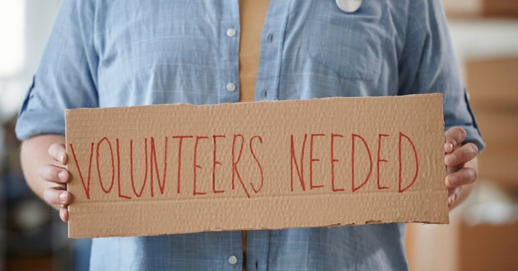 Person holding a sign reading 'Volunteers Needed' to encourage community support.